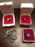 Top view of all four jewelry pieces in three display cases on a wooden surface showing two rings in white cases with red velvet, a third ring in a smaller box with a dark red interior, and a brooch placed on the table.