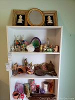 Wide view of white shelving unit with over 40 pieces including Easter bunnies, brass plates, candles, and figurines arranged on three shelves
