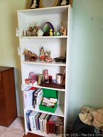 White pressed wood bookshelf filled with decorative figurines, glassware, and books, placed next to a wooden cabinet.