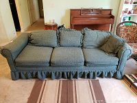 Front view of green upholstered vintage sofa with ruffled skirt on carpeted floor, piano and furniture in background.