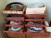 Photo of 10 collectible plates in stacked original boxes, some plates visible showing detailed artwork including winter scenes and landscapes.
