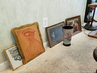 Photo showing multiple framed wall art pieces leaning against a wall, including a portrait sketch in a decorative frame, a small architectural ink drawing, and a tin vase with a painted floral design.