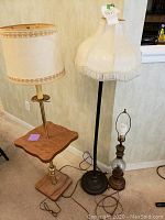 Three lamps: floor lamp with built-in table and decorated beige shade, floor lamp with black stand and mismatched cream shade with fringe, small oil lamp style table lamp without bulb or shade