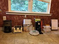 Single image showing the group of kitchen appliances and accessories on the floor: two toasters, coffee maker, slicer, grilling fork, simmer server, wire shelf rack, pots, and a framed item.