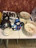 Full lot showing stacked floral patterned Mikasa Heritage Samantha dinnerware plates and bowls, assorted stainless steel flatware in white tray, stainless steel tea kettle with black handle, snowman teapot, and sugar bowl.