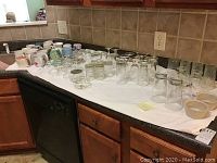 Wide view of over 70 assorted glasses, mugs, and two Corningware bowls laid out on a kitchen countertop.