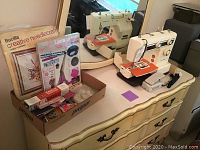 Image showing New Home sewing machine, instruction manual, foot pedal, and a box of assorted sewing and needlecraft items on a dresser.