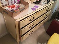 Front and side view of cream-colored French Provincial style dresser with three drawers, showing ornate drawer handles and scalloped detail on bottom edge.
