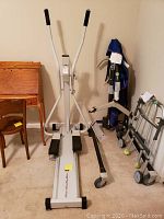 Front view of white and black Skimaster Welder XC-1 exerciser machine with foot pedals and handles, placed indoors near a chair and walker.