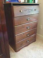 Full view of a medium brown wooden chest of drawers with five drawers, showing top drawer decorative carving and mixed handles.