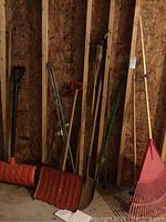 Photo showing yard tools including a red shovel, metal pointed shovel, and edger with long wooden handles leaning against a wood wall in a basement.