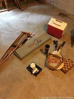 Photo showing items on floor including wooden crutches, boxed Spalding tennis racket in protective frame, Igloo Playmate cooler, chess set, and canister of tennis balls.