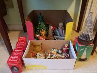 Box with assorted Christmas tree ornaments including small Santa figurines, green tree decoration, and others.