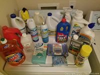 Overview of assorted laundry and cleaning products arranged on a surface, showing various bottles of detergent, bleach, spray cleaners, dryer sheets, and fabric scrubbing products.