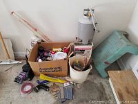 Wide view showing a cardboard box filled with assorted pest control products, hand tools, and garden gloves next to a rusted garden trowel and a large metal tank sprayer with hose.