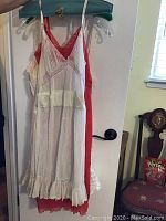Photo showing white slip with ruffled hem, red nightgown behind it, hanging on clear plastic hangers