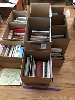 Ten boxes filled with assorted cookbooks including some hardcovers and spiral bound books, arranged on a wooden floor.