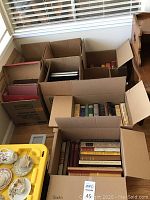 Eight cardboard boxes filled with books arranged on floor near window and yellow container of china