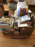 Four boxes and containers with assorted craft supplies, gift wrap, yarn, paper, and scissors arranged on floor.