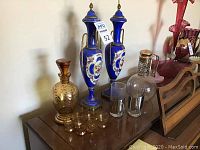 Full lot showing amber gold filigree decanter with matching glasses, two blue painted urns, clear decanter with chrome lid, and two chrome and glass cups on wooden surface.
