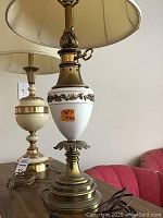 Full view of the two lamps on a wooden table, showing the white ceramic and brass lamp with leaf garland and the brass ribbed lamp.
