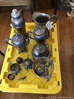 Set of tarnished silver plate serving ware items including teapots, creamers, sugar bowls, cocktail shaker, and other pieces on a yellow bin.