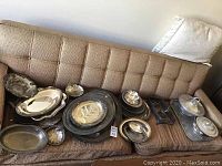 Overview of 30 pieces silver plated serving ware on brown couch including trays, platters, bowls, lidded serving dish, salt and pepper holder.