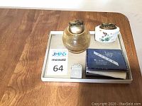 Four vintage lighters displayed on a metal tray, showing the round brass lighter, white floral ceramic lighter, and instruction booklet.