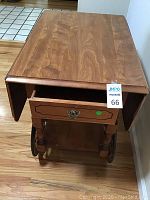 Front view of vintage wooden rolling serving cart showing drop side extensions folded down, single drawer open, spindle legs and wooden wheels with spokes.