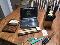 Set of steak knives in metal tray, wooden knife block with Faberware knives, boxed Carvel Hall by Chas. D Briddell knives, paring knives, sharpening tools and skewers on wooden table.