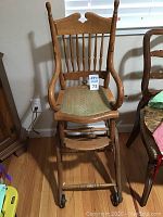 Front view of antique wooden high chair showing woven seat, spindle back, rolling casters, and broken arm of tray.