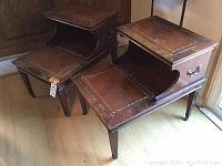 Pair of vintage wooden wheeled side tables with inset bordered tops, drawer, curved open shelf, four legs and caster wheels, placed on wooden floor near window