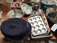 Group of various bakeware and cookware items including cake molds, roasting pan, pots, pans, and kettles arranged on a table showing usage wear.