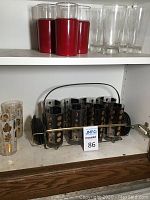 Shelf showing six black glasses with gold dots in a metal carrying tray, two clear glasses with gold leaf designs, and other glasses nearby