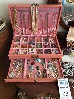 Open pink jewelry chest showing multi-compartment tray with assorted earrings, brooches and necklaces draped over lid