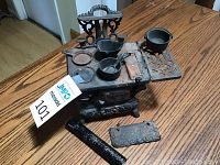 Photo of vintage Crescent cast iron toy oven with cooking accessories including pot, pan, and detachable side panel on wooden table