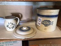 Front view of the crock with lid and small floral pitcher on wooden shelf