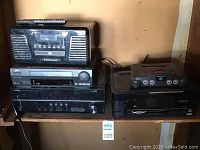 Front view of Crosley CD player stereo on top, JVC VHS player and Yamaha RX-V373 receiver below, Nintendo 64 console on adjacent shelf with Epson printer.
