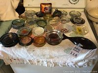 Overall view of the collection of approximately 29 vintage ashtrays spread on a towel on a stove top, showing a variety of shapes, sizes, colors, and materials.