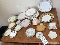 Overview of approximately 40 mismatched vintage plates and decorative dishware items arranged on a wooden table.