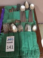 Green fabric roll-up holder with multiple pockets showing silverplate flatware including knives, forks, spoons, and serving pieces arranged in sets.
