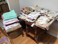 Large gathering of folded tablecloths, napkins, doilies, and tea towels on and around a wooden serving cart and chair.
