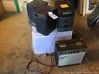 Wide view showing Squire guitar amplifier, JVC portable stereo main unit, and one detached speaker on a white plastic bin in a garage space.