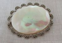 Front view of the oval mother of pearl brooch with scalloped marcasite stone frame on textured white fabric.