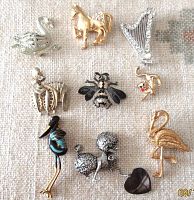 Photo showing 9 vintage themed brooches including animal shapes, a musical instrument, a clown face, and a jester face on a fabric background.