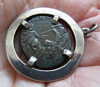 Front side of the Roman coin showing a scene with figures, mounted in a silver frame pendant held in hand.
