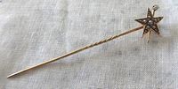 Long view of gold stick pin with star head placed on white fabric background showing the twisted stick shaft and star design with pearls and a small diamond.