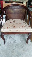 Front view of wooden chair showing curved arms, cane backrest, and floral cushioned seat on concrete floor.