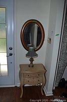 Photo showing wood framed oval mirror mounted on wall, small distressed wooden stand table beneath it, and small decorative lamp placed on table.