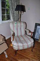 Front diagonal view of upholstered wingback chair showing striped fabric and wooden armrests and legs, lamp behind.